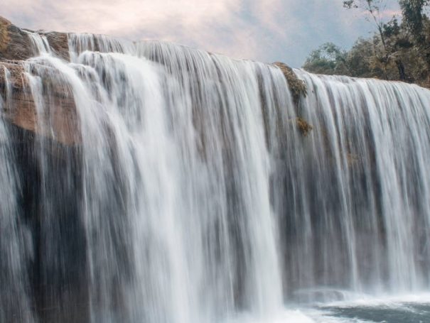 pexels-photo-3653963-3653963-2 Peaceful waterfall cascading over rocks with lush forest backdrop and serene sky.